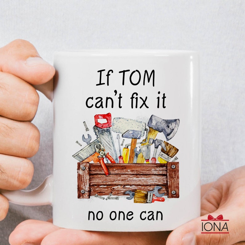 May include: White ceramic mug with the text "If TOM can't fix it, no one can." The mug features a watercolor illustration of a wooden toolbox filled with various tools, including a saw, hammer, and wrench.
