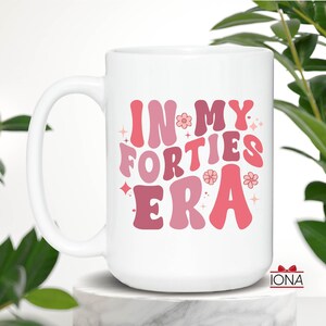 May include: White ceramic mug with the text "IN MY FORTIES ERA" in shades of pink and red. The mug has a large handle and is decorated with flowers and stars. The mug is a perfect gift for someone celebrating their 40th birthday.