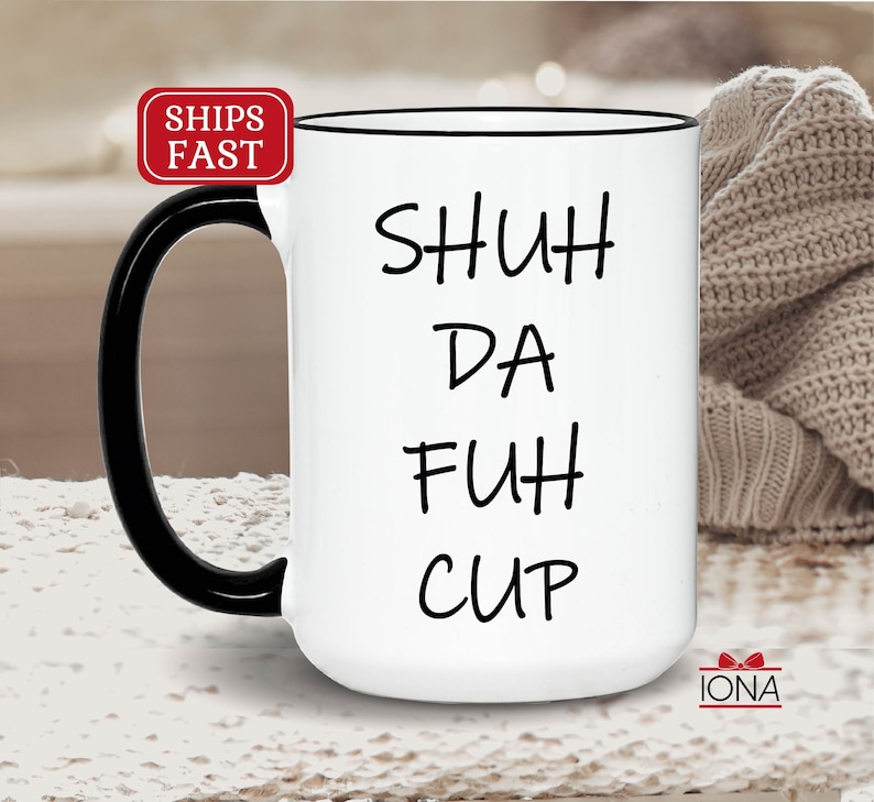 K&ouml;nnte beinhalten: Wei&szlig;e Keramik-Kaffeetasse mit schwarzem Henkel und schwarzem Rand. Die Tasse tr&auml;gt den Schriftzug "SHUH DA FUH CUP" in Schwarz.