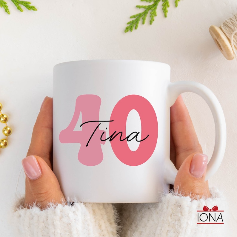 May include: White ceramic mug with a pink "40" and the name "Tina" in a script font. The mug is held by a person wearing a white sweater. A gold beaded necklace and greenery are in the background.