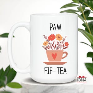 May include: White ceramic mug with a black handle. The mug has a pink teacup with a red heart and flowers in it. The text "PAM FIF-TEA" is printed on the mug.
