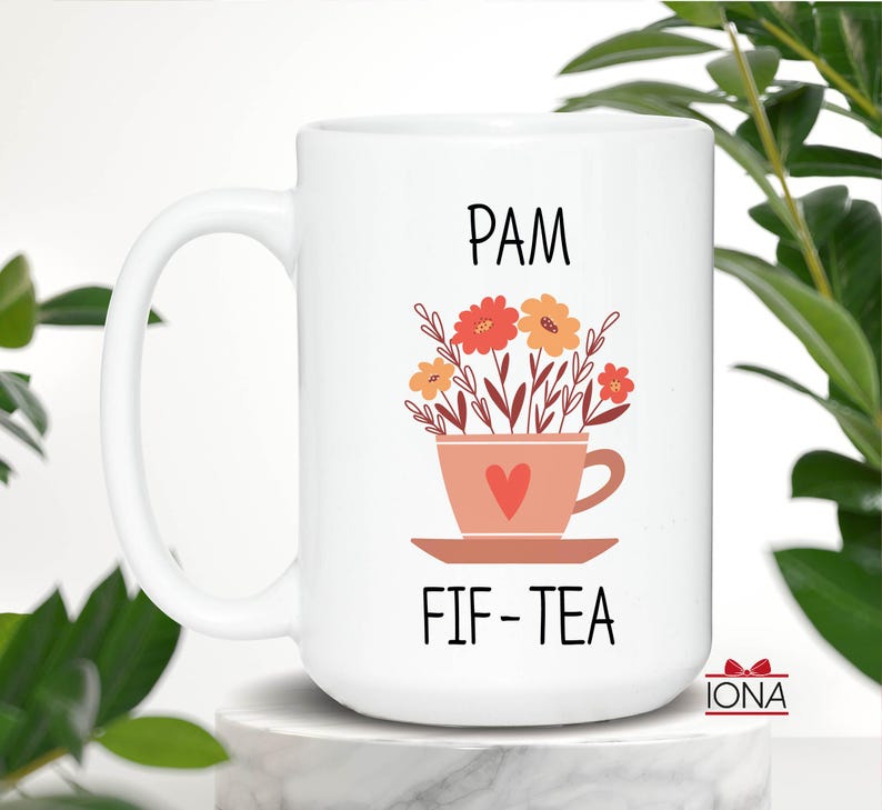 May include: White ceramic mug with a black handle. The mug has a pink teacup with a red heart and flowers in it. The text "PAM FIF-TEA" is printed on the mug.