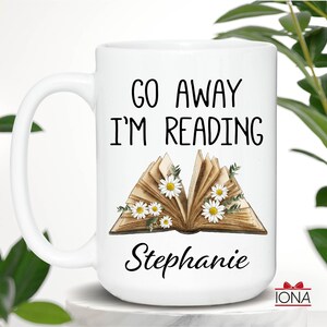 Op de afbeelding: Een witte keramische mok met een zwart handvat. De mok heeft de tekst "Go Away I'm Reading" en een bloemige illustratie van een open boek met madeliefjes. De naam "Stephanie" staat onder het boek.