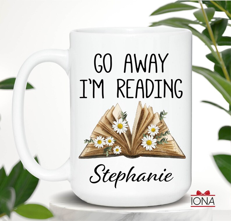 Op de afbeelding: Een witte keramische mok met een zwart handvat. De mok heeft de tekst "Go Away I'm Reading" en een bloemige illustratie van een open boek met madeliefjes. De naam "Stephanie" staat onder het boek.