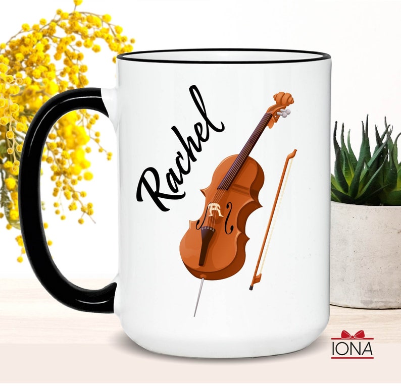 May include: White ceramic mug with a black handle and black rim. The mug has a cello and bow illustration and the name "Rachel" written in black cursive.