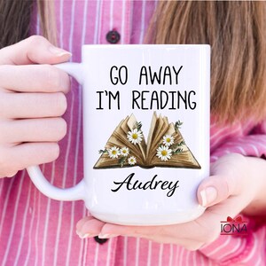 Op de afbeelding: Witte keramische mok met de tekst "Go Away I'm Reading" en een gestileerde afbeelding van een open boek met witte bloemen. De naam "Audrey" staat onder het boek gedrukt.