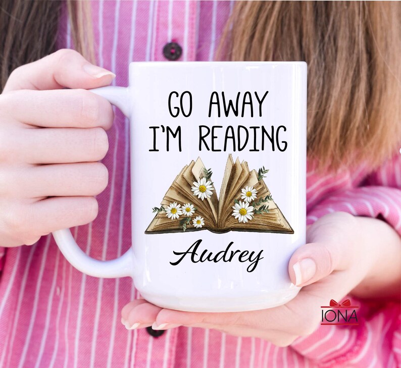 Op de afbeelding: Witte keramische mok met de tekst "Go Away I'm Reading" en een gestileerde afbeelding van een open boek met witte bloemen. De naam "Audrey" staat onder het boek gedrukt.