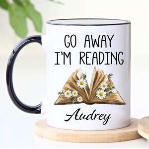 Op de afbeelding: Witte keramische mok met een zwarte rand en handvat. De mok heeft een zwarte afbeelding met de tekst "Go Away I'm Reading" met een vintage boekillustratie en de naam "Audrey".