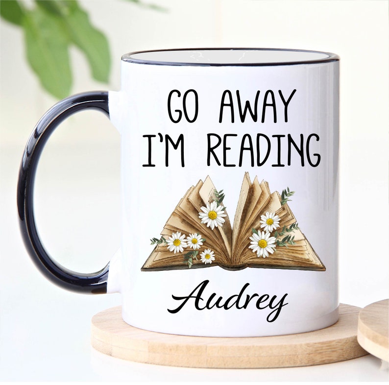 Op de afbeelding: Witte keramische mok met een zwarte rand en handvat. De mok heeft een zwarte afbeelding met de tekst "Go Away I'm Reading" met een vintage boekillustratie en de naam "Audrey".