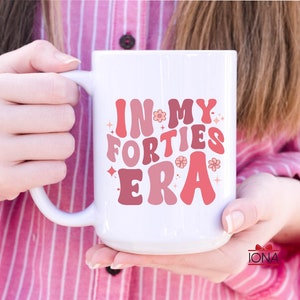 May include: White ceramic mug with the text "IN MY FORTIES ERA" in shades of pink and red. The mug features a curved handle and is decorated with floral and star accents. A fun and stylish gift.