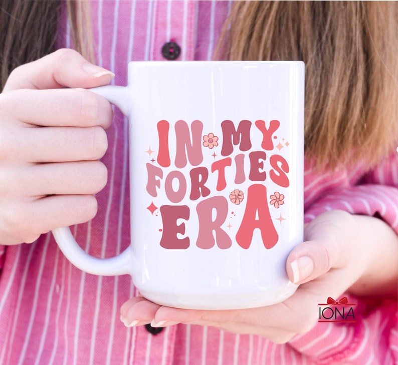 May include: White ceramic mug with the text "IN MY FORTIES ERA" in shades of pink and red. The mug features a curved handle and is decorated with floral and star accents. A fun and stylish gift.