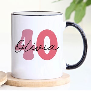 May include: White ceramic mug with a black handle and rim. The mug features the number "40" in a pink hue, with the name "Olivia" in a cursive font. The mug sits on a wooden coaster.