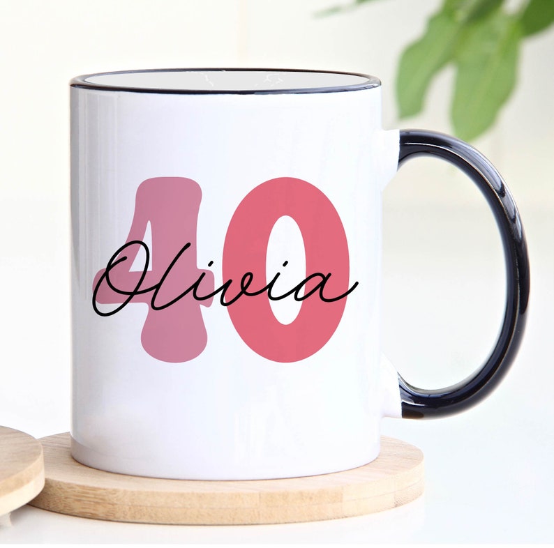 May include: White ceramic mug with a black handle and rim. The mug features the number "40" in a pink hue, with the name "Olivia" in a cursive font. The mug sits on a wooden coaster.