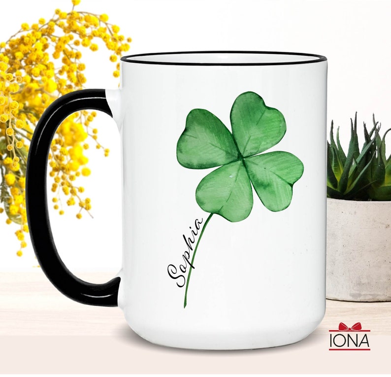 May include: White ceramic mug with a black handle and rim. The mug features a green four-leaf clover with a stem and the name "Sophia" written in black cursive.