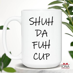 K&ouml;nnte beinhalten: Wei&szlig;e Keramik-Kaffeetasse mit schwarzem Text, der "SHUH DA FUH CUP" lautet.