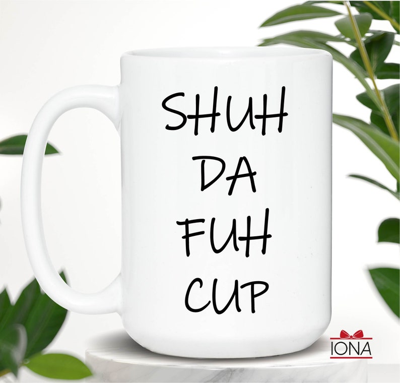 K&ouml;nnte beinhalten: Wei&szlig;e Keramik-Kaffeetasse mit schwarzem Text, der "SHUH DA FUH CUP" lautet.