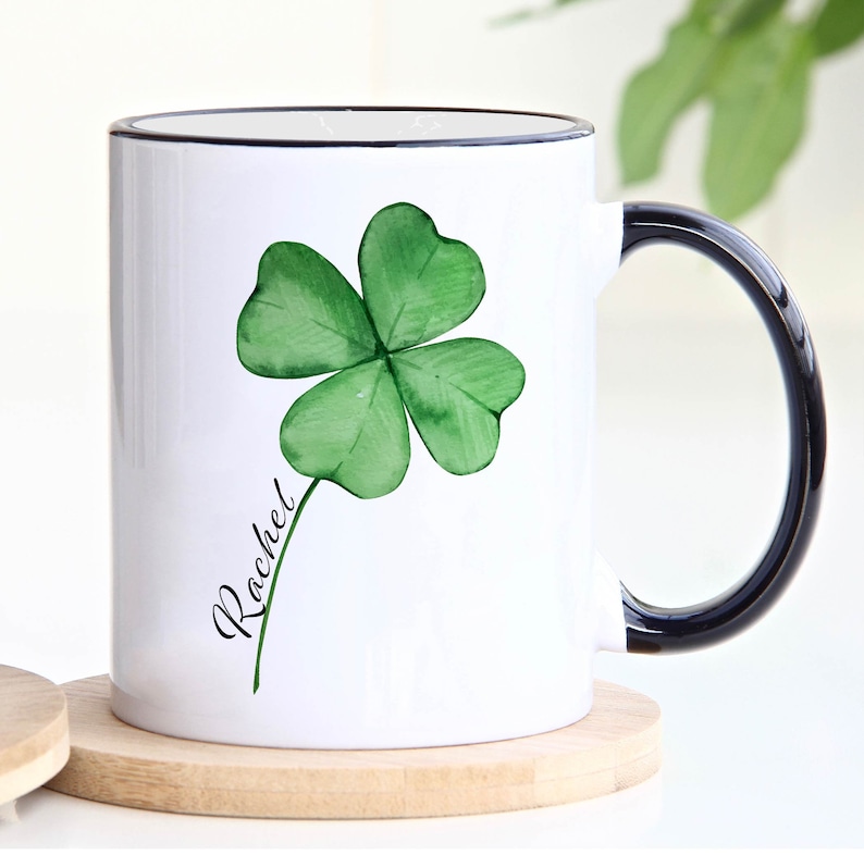 May include: White ceramic mug with a black rim and handle. The mug features a green four-leaf clover with a stem and the name "Rachel" written in cursive.