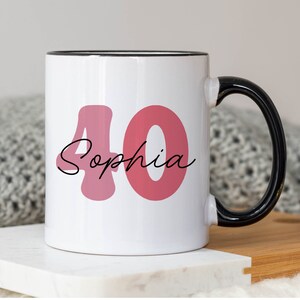 May include: White ceramic mug with a black handle and rim. The mug features the number "40" in a pink hue, with the name "Sophia" in a cursive font. The mug is designed for a birthday celebration.