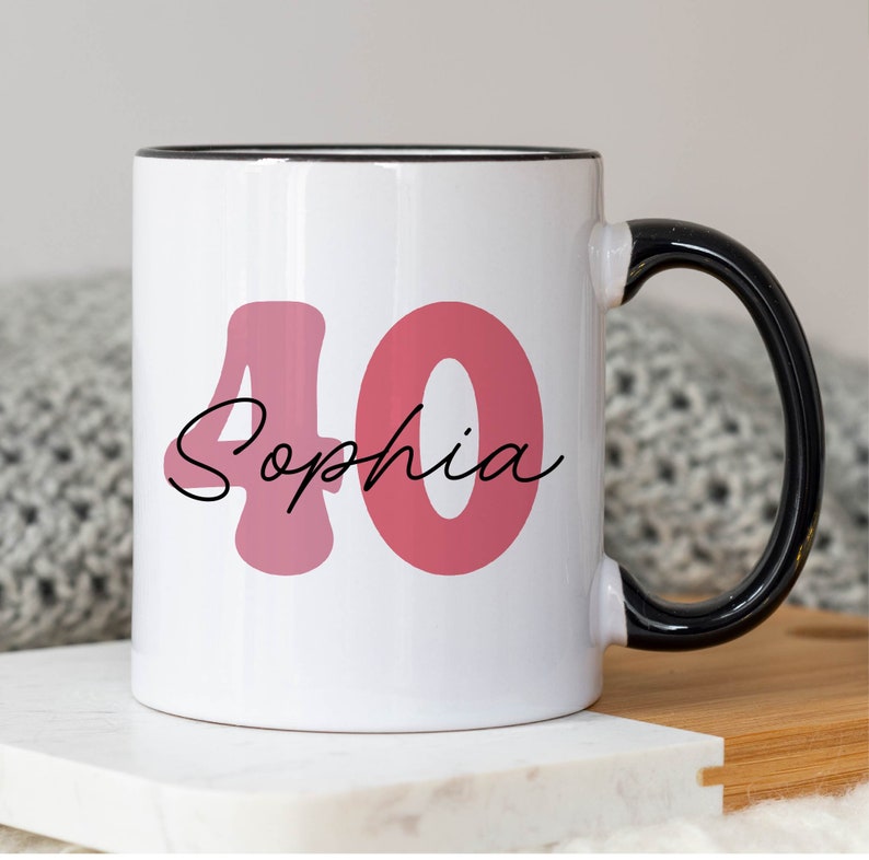 May include: White ceramic mug with a black handle and rim. The mug features the number "40" in a pink hue, with the name "Sophia" in a cursive font. The mug is designed for a birthday celebration.