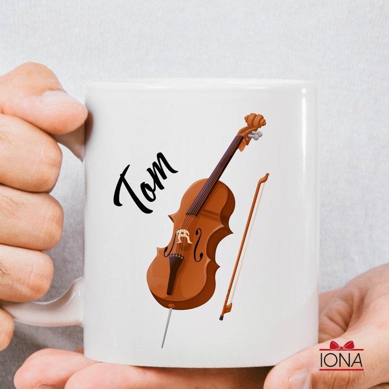 May include: White ceramic mug with a black and brown illustration of a cello and the name "Tom" printed on the side.