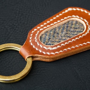 May include: A handcrafted leather keychain with a brass ring. The brown leather has white stitching around a central oval of blue and tan tweed fabric. The keychain is designed for holding keys.