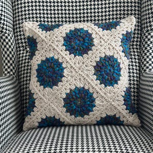 May include: A square crochet pillow featuring a repeating pattern of cream-colored yarn squares joined together. Each square contains a circular floral motif worked in variegated shades of deep blue, teal, and purple yarn with a textured stitch.