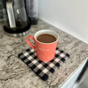 May include: A coral-colored mug filled with coffee sits on a black, white, and gray checkered crochet coaster. The mug has a white interior and the phrase "c'est la vie" printed on the side. The mug is on a granite countertop.