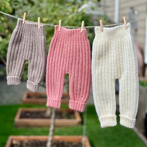 May include: Three pairs of handmade baby pants in varying colors hang on a clothesline. The pants are crocheted in a ribbed pattern. Colors include a light taupe, a soft pink, and a creamy white. The pants have a high waist and cuffed ankles.