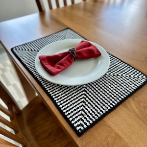 May include: A white plate with a folded red napkin and a black napkin ring sits on a black and white patterned placemat. The placemat is on a wooden table. The table is made of wood and has a glossy finish. The setting is for a meal.