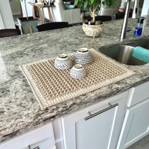 May include: A rectangular beige crochet dish drying mat with a textured waffle stitch pattern. Three small ceramic bowls with black and white geometric patterns are placed on top of the mat, which rests on a granite kitchen countertop.