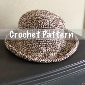 May include: A crocheted hat with a wide brim, featuring a brown and cream color scheme. The hat has a textured appearance and the words "Crochet Pattern" are displayed across the front. The hat is sitting on a dark surface.