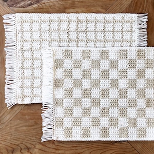 Może przedstawiać: Dwa szydełkowe podkładki pod talerze w kratkę w kolorze beżowym i białym. Podkładki mają białe frędzle wokół krawędzi.