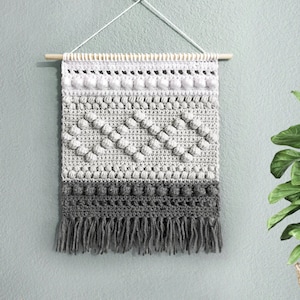 May include: A grey and white crocheted wall hanging with a fringe border. The hanging is made with a variety of stitches and textures, including bobbles and a chevron pattern.