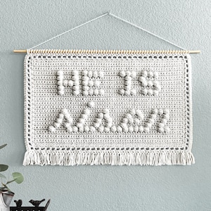 May include: White crocheted wall hanging with the words "Hello" and "Love" in raised letters. The hanging has a fringe border and is hung on a light blue wall.