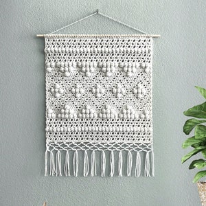 Peut inclure: Suspension murale en macramé blanche avec un motif géométrique et des franges. La suspension est sur un mur vert clair avec une plante en pot et des livres sur une étagère blanche.