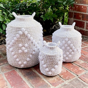 May include: Three crocheted baskets in different sizes, all with a beige base and white pom-poms. The baskets are arranged in a row on a brick surface.