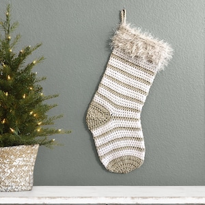 May include: A handmade Christmas stocking with a white and gold striped pattern, hanging against a gray wall. The stocking has a fluffy, light brown trim at the top. A small Christmas tree in a woven basket is to the left.