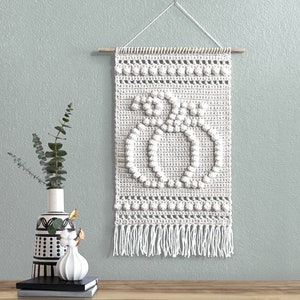 Pode incluir: Decoração de parede de crochê branca com um design de duas abóboras. A decoração tem uma ripa de madeira na parte superior e uma franja na parte inferior.