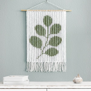 Puede incluir: Un tapiz de pared de crochet blanco con un diseño de hoja verde. El tapiz tiene una varilla de madera en la parte superior y flecos blancos en la parte inferior.