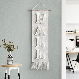 Könnte beinhalten: Weißer gehäkelter Wandbehang mit dem Wort "FALL" in großen Buchstaben aus weißen Pompons. Der Behang ist an einer Holzleiste befestigt und hat unten einen Fransen.