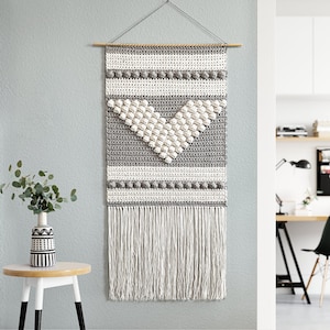 May include: A gray and white crocheted wall hanging with a fringe border. The hanging features a geometric pattern with a heart shape in the center.