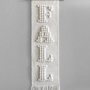 May include: A white crocheted wall hanging with the word "FALL" spelled out in large letters. The letters are made with a bobble stitch and have a 3D effect. The hanging has a fringe at the bottom.