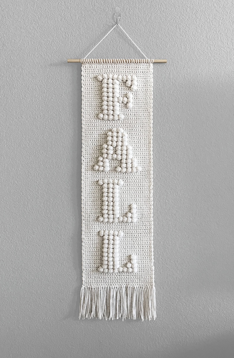 May include: A white crocheted wall hanging with the word "FALL" spelled out in large letters. The letters are made with a bobble stitch and have a 3D effect. The hanging has a fringe at the bottom.