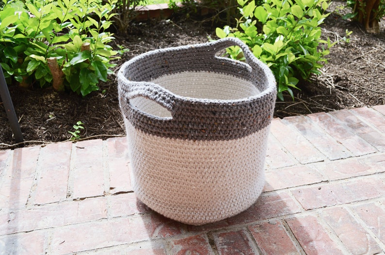 May include: A crocheted storage basket with two handles. The basket is made of white yarn with a grey band around the top.