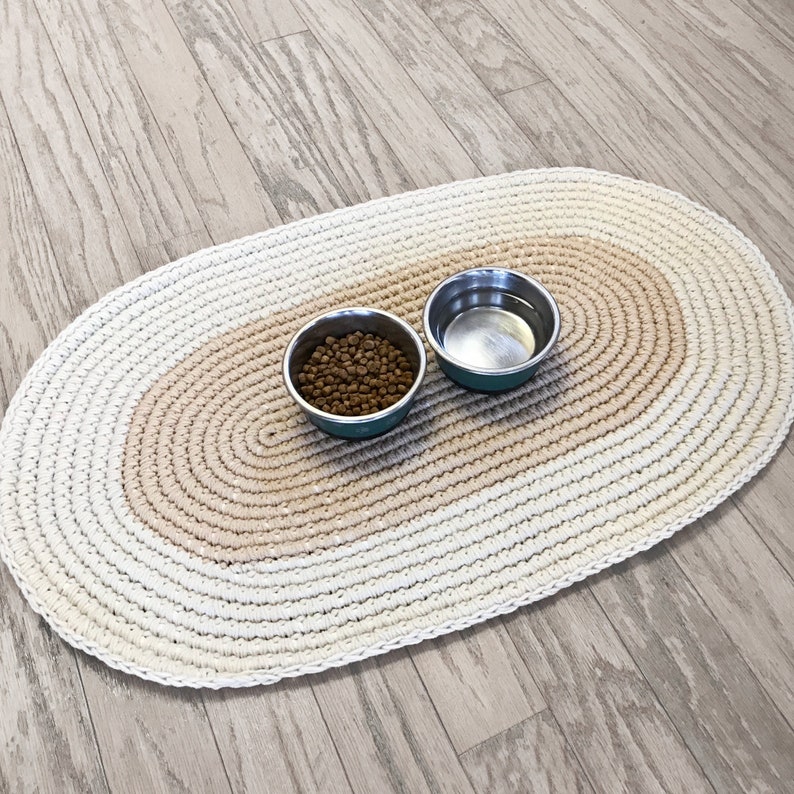 May include: A white oval crochet pet mat with a beige center stripe. Two metal bowls are on the mat, one filled with brown pet food and the other empty.