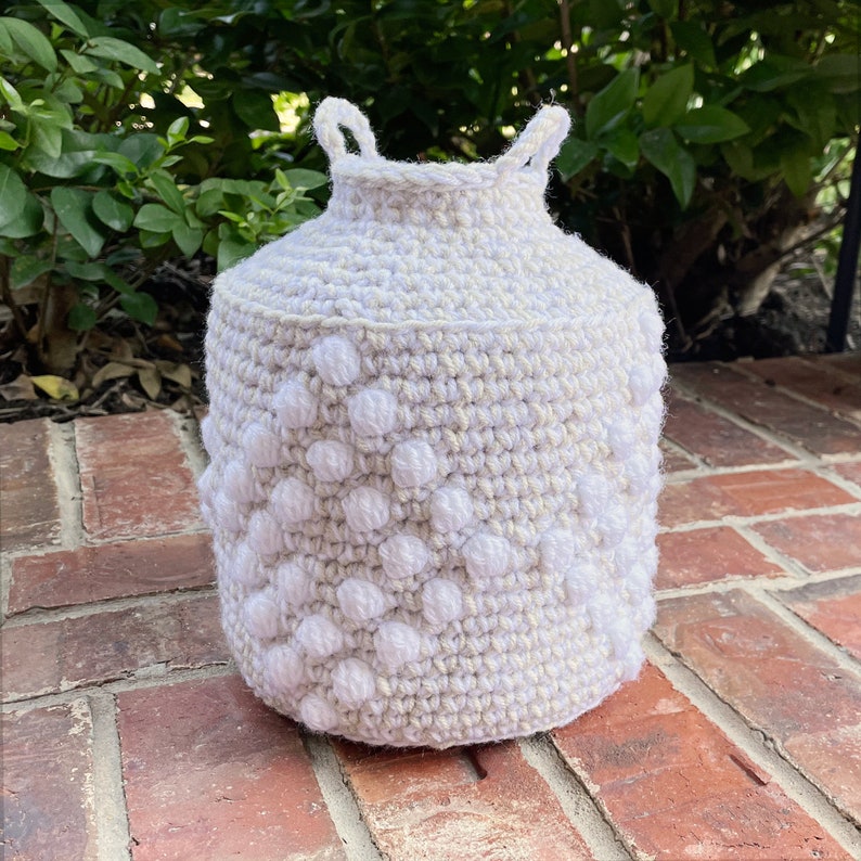 May include: A white crocheted basket with a textured pattern and a handle. The basket is made with white and beige yarn.