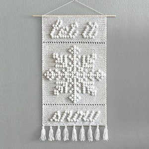 May include: White crocheted wall hanging with a snowflake design and tassels. The snowflake is made of white pom-poms.