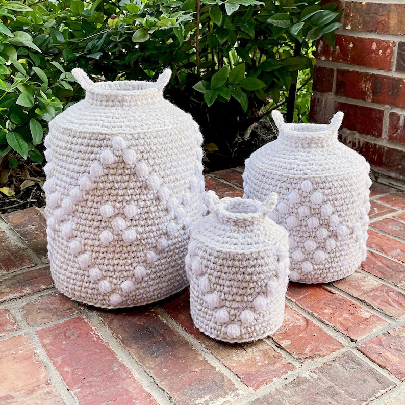 May include: Three crocheted baskets in different sizes, all with a beige base and white pom-poms. The baskets are arranged in a row on a brick surface.