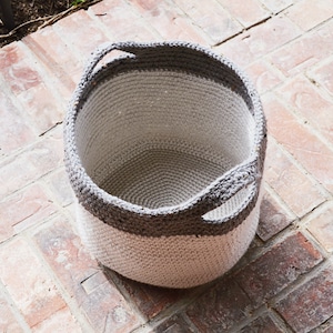 May include: A white and grey crocheted storage basket with two handles. The basket is empty and sitting on a brick surface.
