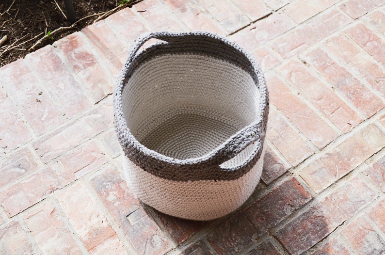 May include: A white and grey crocheted storage basket with two handles. The basket is empty and sitting on a brick surface.